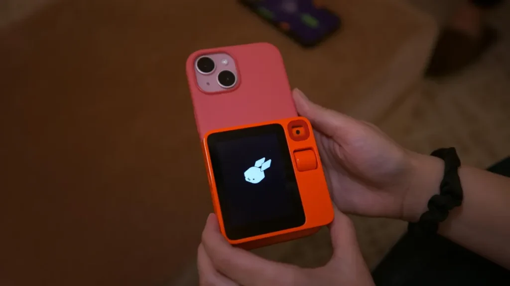 Оранжевый цвет R1, небольшой экран и колесо прокрутки придают устройству яркий и почти ретро стиль. Оно легкое и буквально в полтора раза меньше обычного смартфона, как видно на фотографии R1, расположенной поверх моего iPhone 15. Фактически его форма и размеры напоминают использование раскладного телефона, такого как Samsung Galaxy Z Flip или Motorola Razr 2025, но существенно легче. 